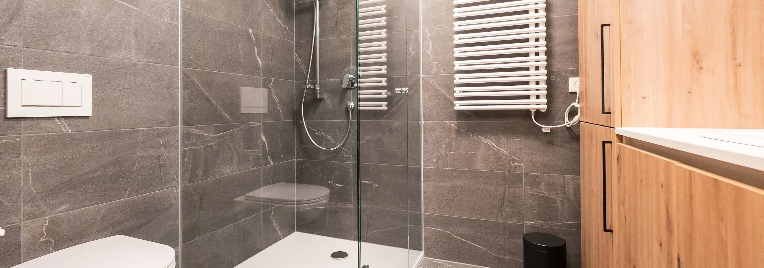 Modernes Badezimmer mit grauen Fliesen, einer ebenerdigen Dusche mit Glastür, hängendem WC, Handtuchheizkörper und Holzwaschtisch mit Waschbecken.