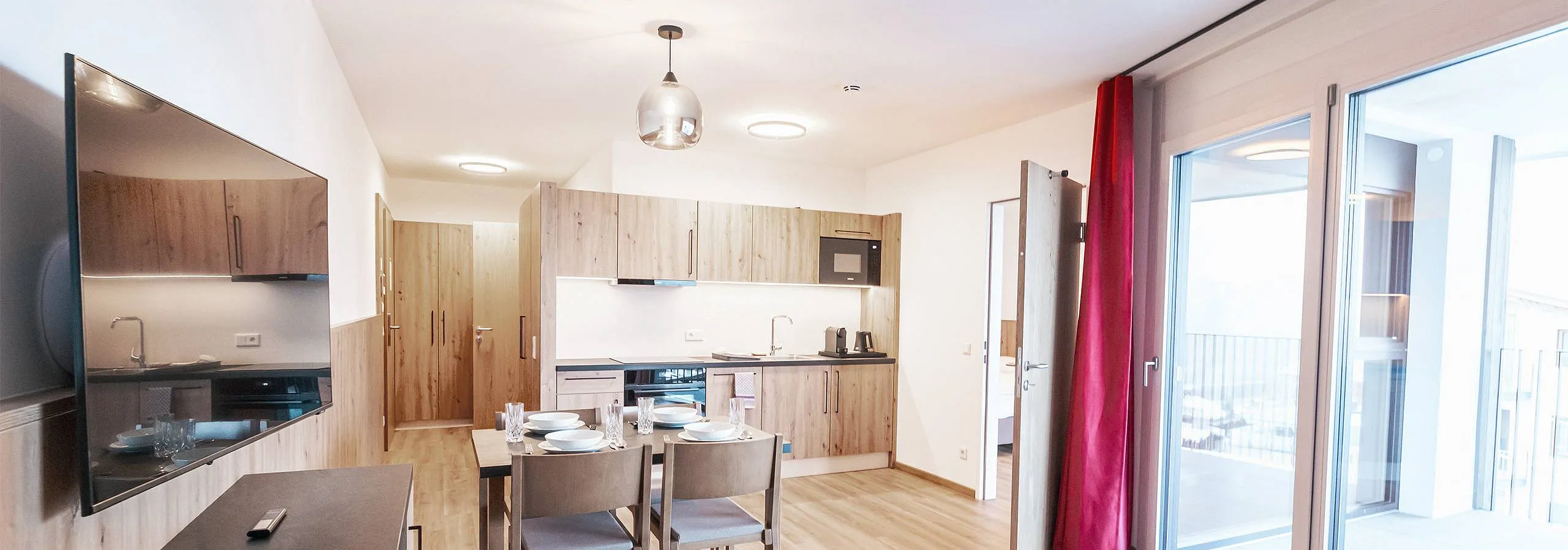 Moderne Küche mit Holzelementen und Esstisch. Der Raum ist hell erleuchtet mit einem hängenden und einem Deckenlicht. Nebenzimmer ist sichtbar.