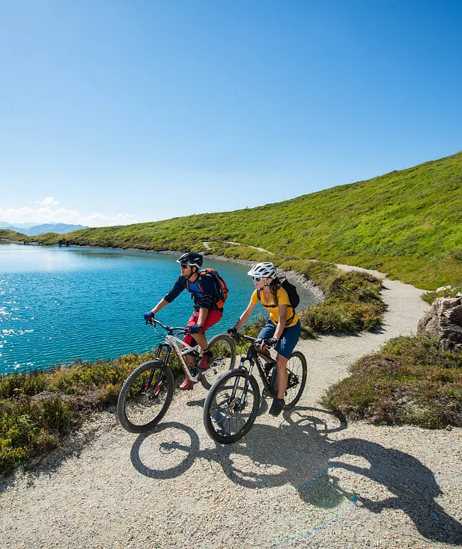 Zwei Mountainbiker fahren auf einem schmalen Pfad entlang eines malerischen Sees in einer grünen Berglandschaft unter strahlend blauem Himmel.