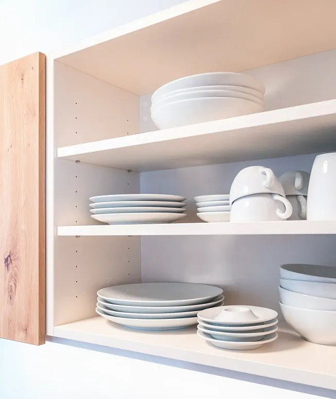 Ein geöffneter Küchenschrank aus Holz zeigt ordentlich gestapelte, weiße Porzellanteller, Schalen und Tassen auf zwei Regalen.