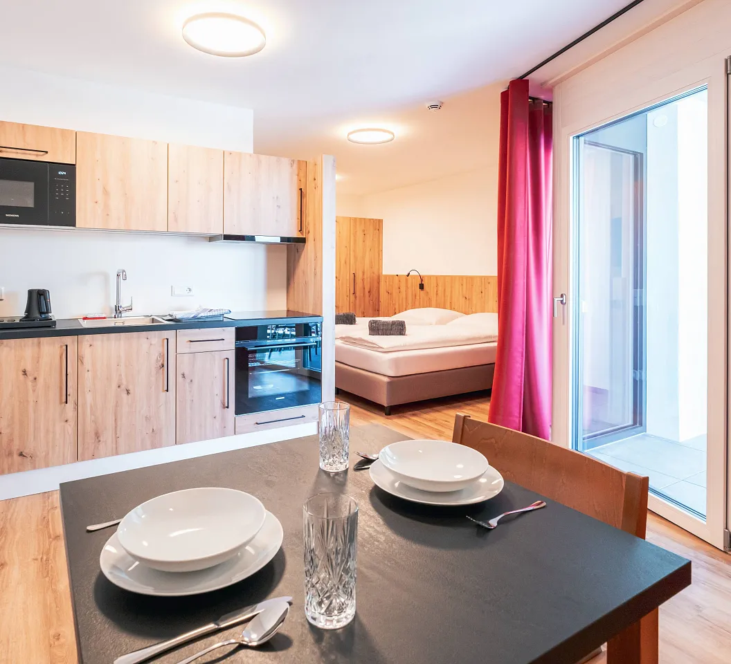 Moderne Einzimmerwohnung mit Holzmöbeln. Offene Küche mit Mikrowelle, Tisch mit Geschirr, Schlafbereich mit Doppelbett, rote Vorhänge und Balkonzugang.