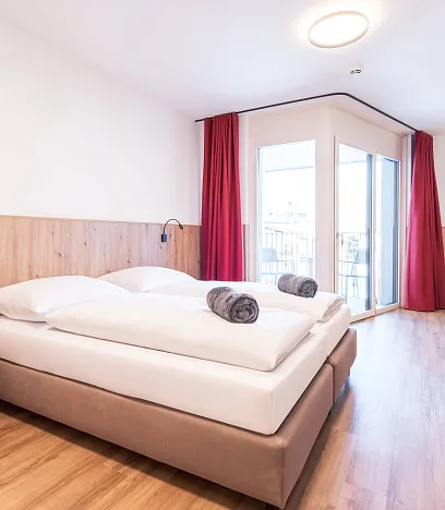 Helles, modernes Hotelzimmer mit Doppelbett, weißen Bettdecken, roten Vorhängen, Holzfußboden und einem Flachbildfernseher an der Wand.