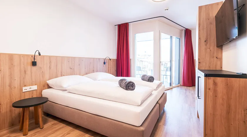 Helles Hotelzimmer mit großem Doppelbett, weißen Bettdecken und Holzverkleidung. Rote Vorhänge, Flachbildfernseher und modernes Dekor. Holzfußboden.