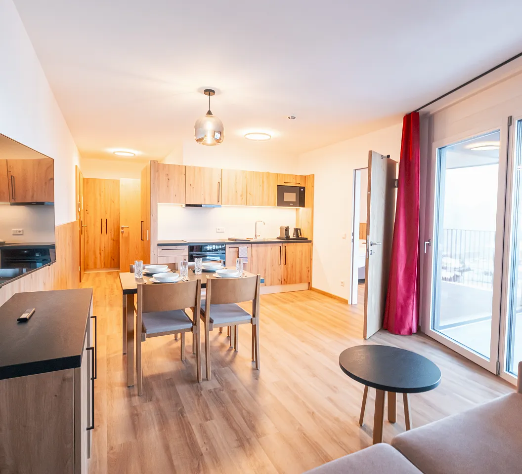 Helles Wohnzimmer mit Holzmöbeln, Esstisch und Stühlen. Eine offene Küche, Flachbildfernseher und große Fenster mit roten Vorhängen sind sichtbar.