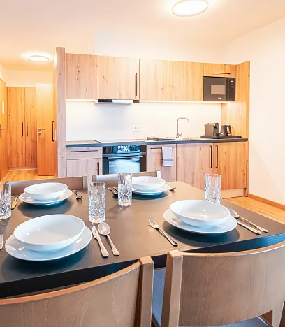 Moderne Küche mit Holzverkleidung, Esstisch mit vier Gedecken, Gläser und Besteck. Im Hintergrund ein Schlafzimmer. Helle, warme Beleuchtung.