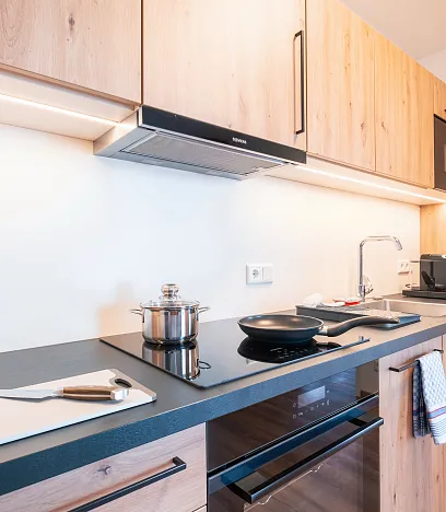 Moderne, schmale Küche mit Holzfronten. Links ein Induktionsherd, darauf ein Topf und eine Pfanne. Oberschränke und eine Mikrowelle darüber, rechts eine Spüle.