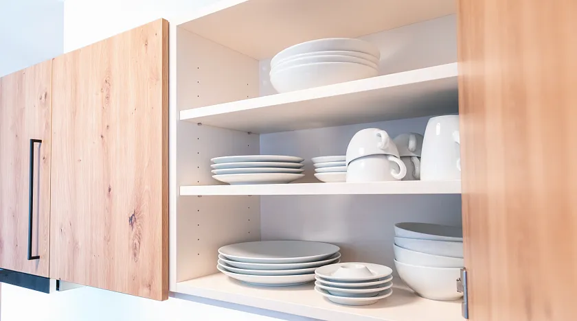 Ein geöffneter Küchenschrank aus Holz zeigt ordentlich gestapelte, weiße Porzellanteller, Schalen und Tassen auf zwei Regalen.