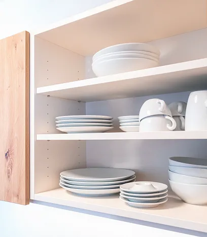 Ein geöffneter Küchenschrank mit zwei Regalböden, auf denen weißes Geschirr, darunter Teller, Schalen und Tassen, ordentlich gestapelt ist.