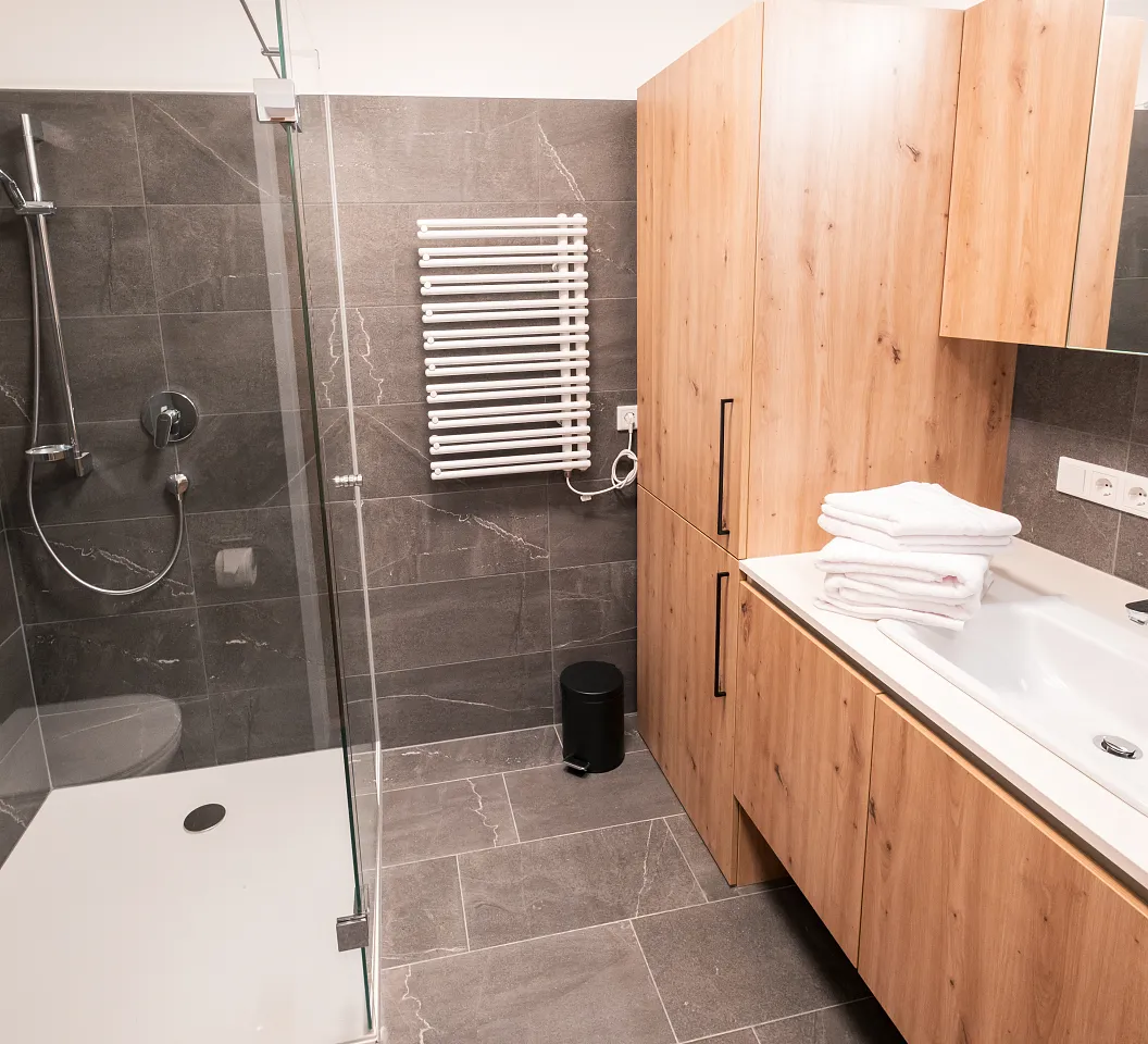 Modernes Badezimmer mit ebenerdiger Dusche, Holzschränken, großem Spiegel und Handtuchheizkörper. Die Wände und der Boden sind aus grauen Fliesen.
