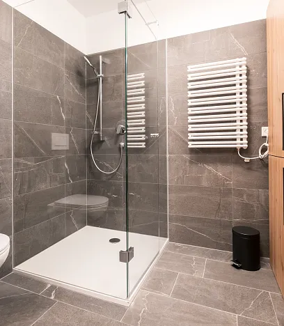 Modernes Badezimmer mit grauen Fliesen, einer Glasdusche, einem hängenden WC und einem Holzschrank. Handtuchheizkörper und Mülleimer sind ebenfalls zu sehen.