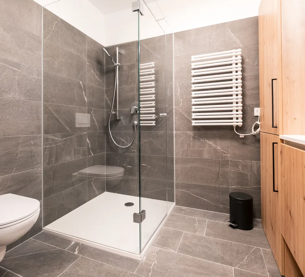Modernes Badezimmer mit grauen Fliesen, Glasdusche, Wandtoilette und Holzschrank. Handtuchheizkörper an der Wand, minimalistisches Design.