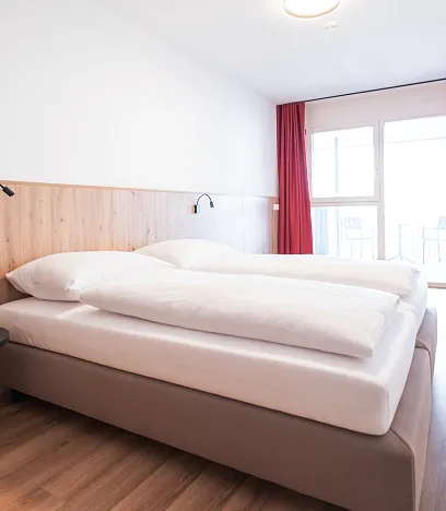 Helles Schlafzimmer mit Holzfußboden und Holzvertäfelung an der Wand. Ein großes Bett mit weißer Bettwäsche steht neben einem kleinen runden Nachttisch.