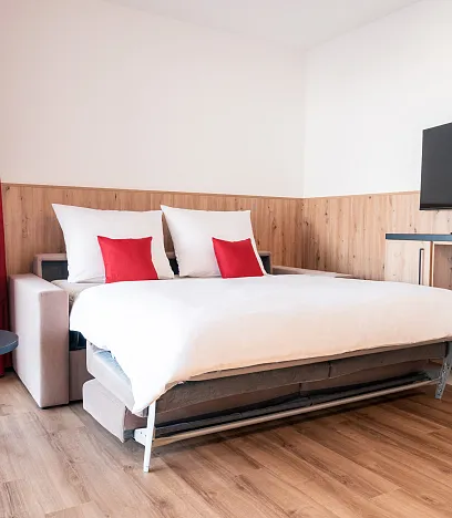 Modernes Hotelzimmer mit einem Doppelbett, weißen Bettdecken, roten Kissen und Holzwänden. Ein Flachbildfernseher steht auf einem schlichten Tisch.