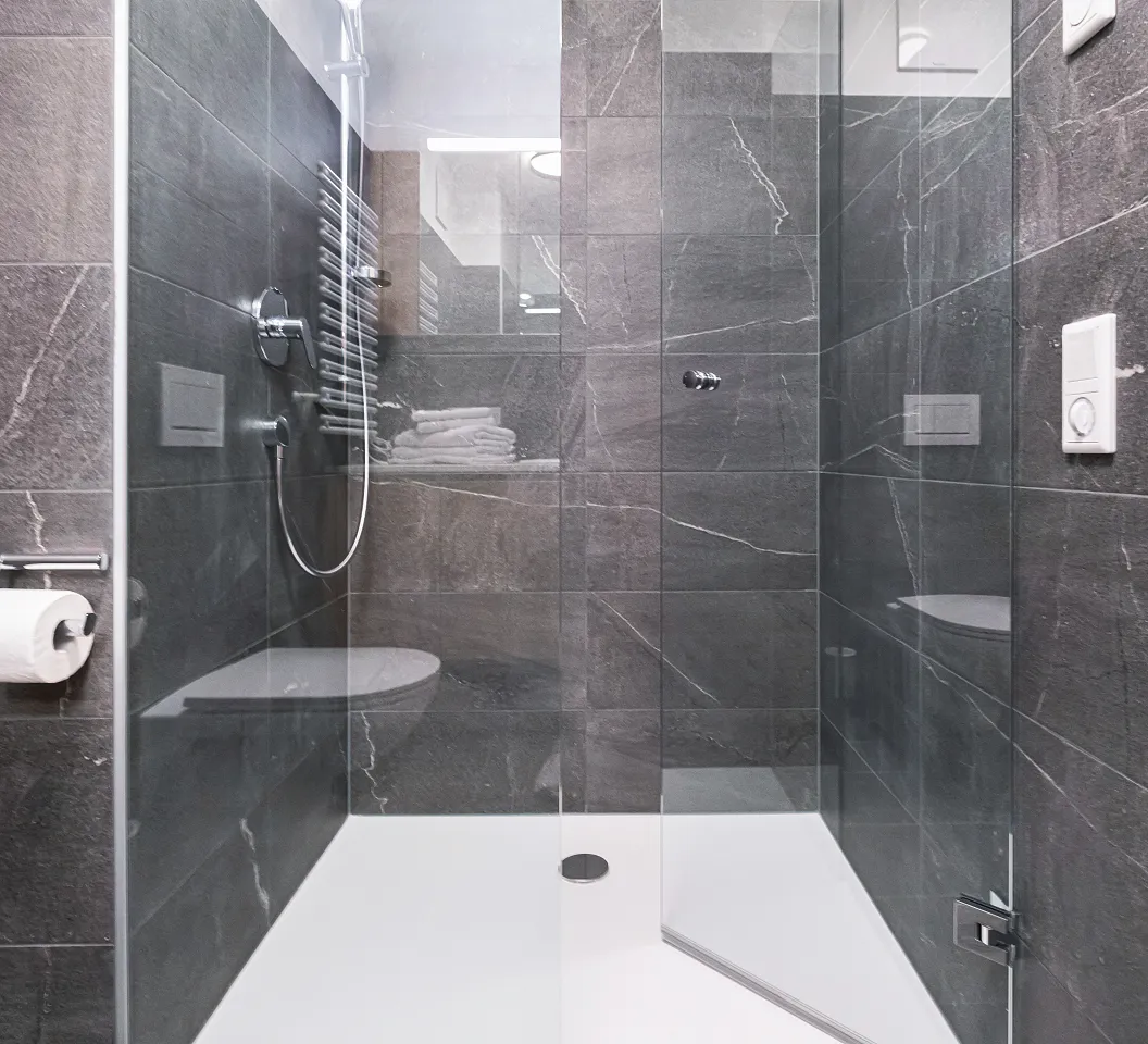 Moderne, minimalistische Duschkabine mit dunklen Fliesen, Glastüren, einem Regenduschkopf und weißen Sanitärutensilien. Ein Handtuch ist sichtbar.