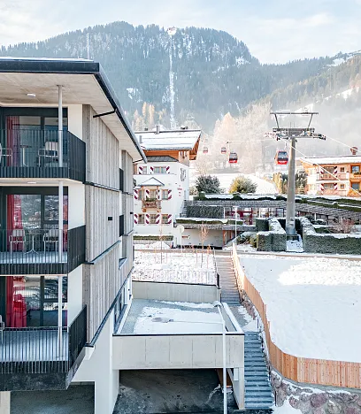 Moderne Bergresidenz mit Holzverkleidung und Balkonen, nahe einer Seilbahnstation in schneebedeckter, bergiger Landschaft.