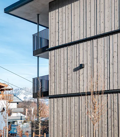 Moderne Holzwand eines Gebäudes mit Balkon, eine rote Seilbahnkabine fährt im Vordergrund. Im Hintergrund sind schneebedeckte Berge unter blauem Himmel.