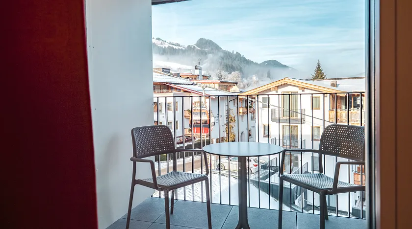 Blick von einem Balkon mit zwei leeren Stühlen und einem runden Tisch auf schneebedeckte Berge und Häuser in einem winterlichen Dorf.
