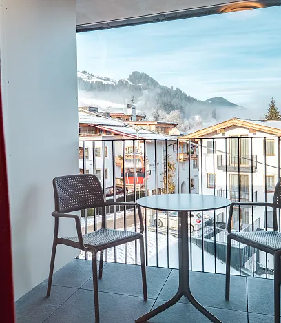 Ein Balkon mit zwei leeren Stühlen und einem Tisch bietet einen Blick auf schneebedeckte Berge und ein Dorf bei klarem Himmel.