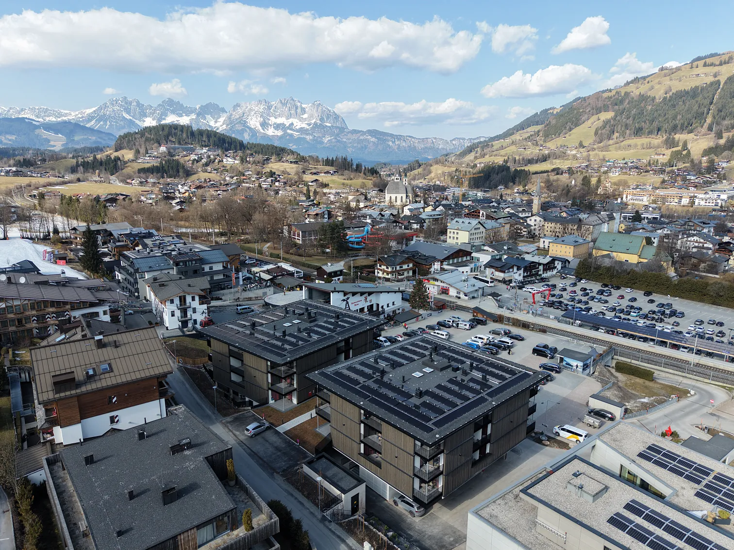 Luftaufnahme einer alpenländischen Stadt mit Bergkulisse. Moderne Wohngebäude im Vordergrund, traditionelle Häuser und schneebedeckte Berge im Hintergrund.