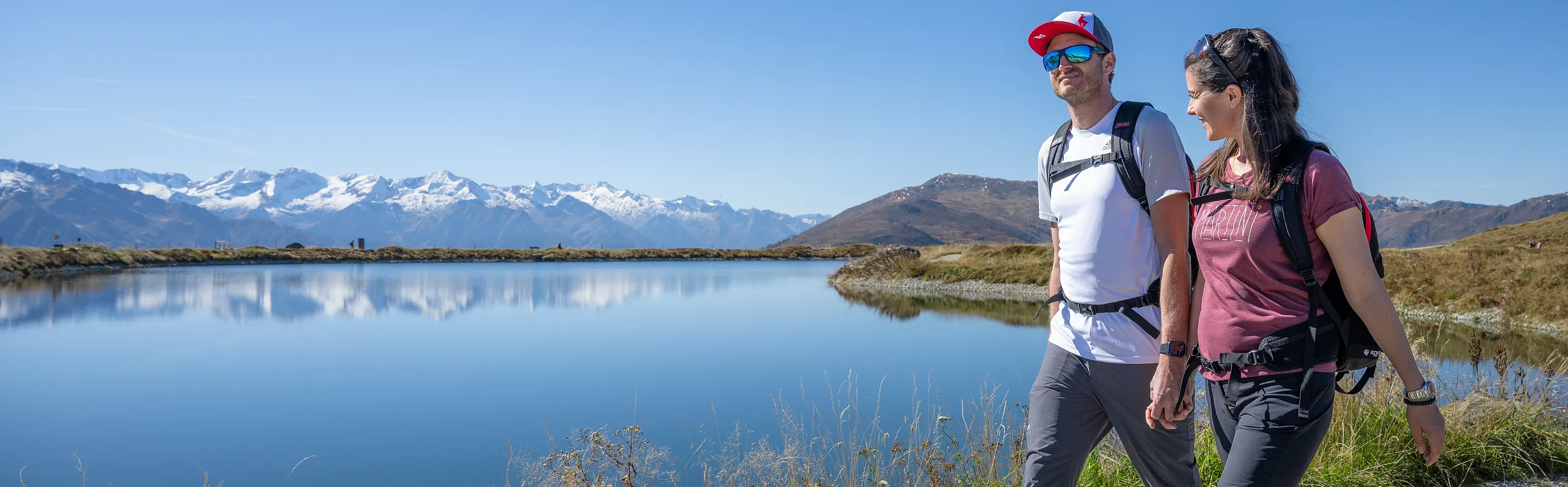 Ein Wanderer mit Sonnenbrille und roter Kappe steht vor einem klaren Bergsee. Im Hintergrund schneebedeckte Berge und blauer Himmel.