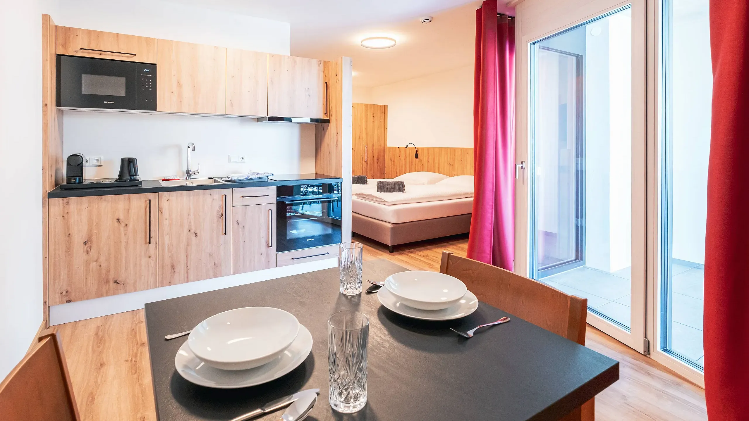 Moderne Wohnung mit offener Küche, Esstisch und Schlafzimmer. Holzmöbel, große Fenster mit roten Vorhängen, gemütliche und helle Atmosphäre.