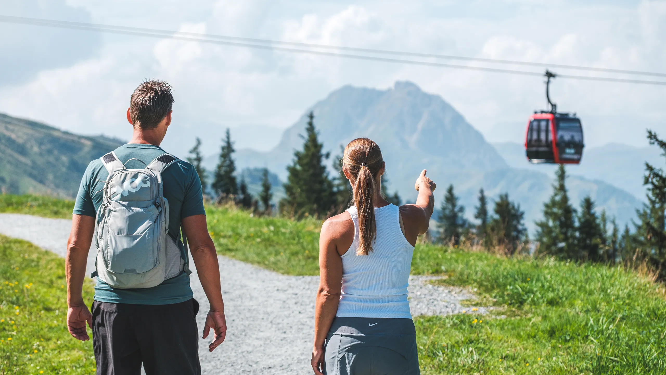 Ein Mann und eine Frau wandern auf einem Bergpfad mit Blick auf eine Berglandschaft. Die Frau zeigt auf eine Seilbahn, die vor einem großen Berg vorbeifährt.